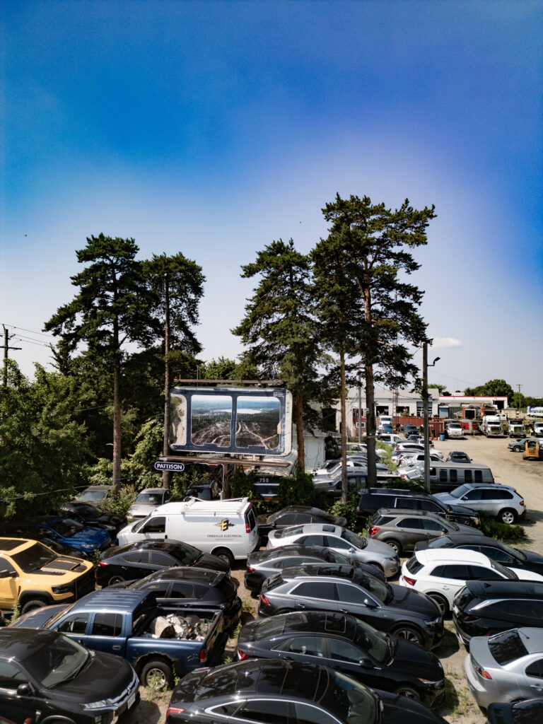 A colour photograph of a Pattison billboard of a photomontage of two photographs. The billboard is surrounded by trees and above a lot of cars parked closely together.
