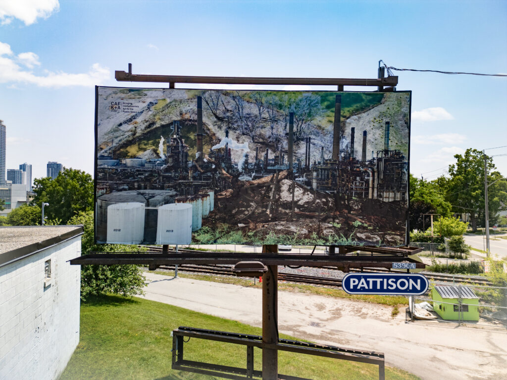 A colour photo of a billboard featuring a photo montage of factory images, smoke stacks, burned trees and a landscape. The billboard is in an urban landscape surrounded by buildings.
