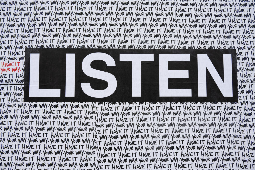 Colour Photograph of art pasted up on a black wall. The word LISTEN in white text on black and the words "have it your way" repeated in the background.