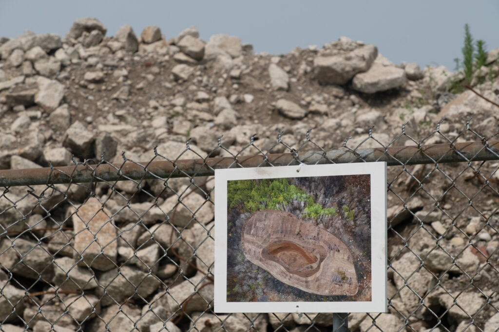 A photograph of a paste up collage that looks like a pit surrounded by burned forest on a white board affixed to a chainlink fence. Behind the fence is a pile of stone rubble,