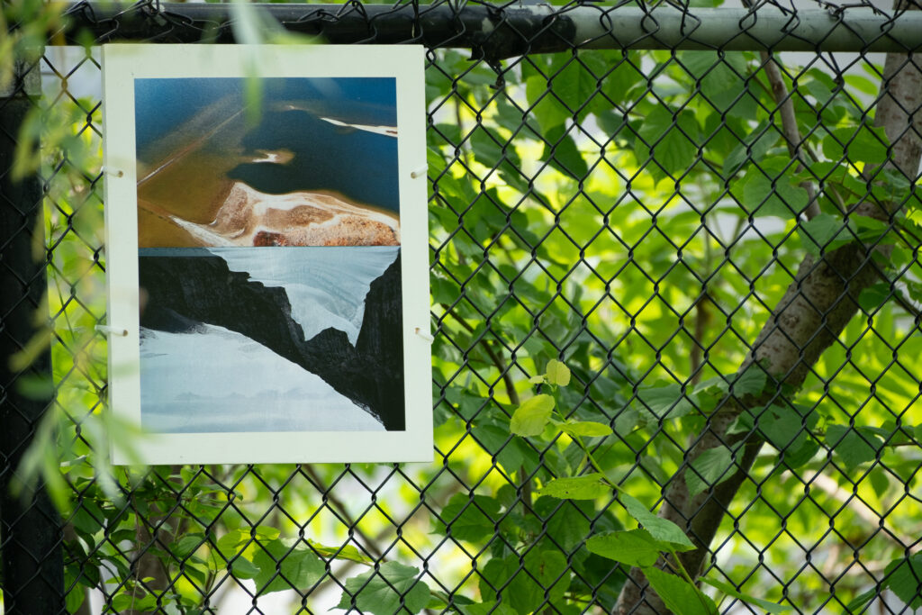 A clour photograph of a photomontage of two images joined at the horizontal center. The top is an aerial image of a lake bottom of sand and gravel, a submerged old roadway and wall can be seen. the bottom image is of an iceberg and mountain range. The image shapes (land and iceberg) mirror each other. The plaque is attached to a chain link fence. Leafed out tree branches surround the plaque.