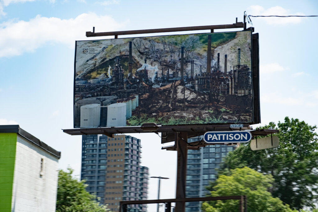 A colour photo of a billboard featuring a photo montage of factory images, smoke stacks, burned trees and a landscape. The billboard is in an urban landscape surrounded by buildings.