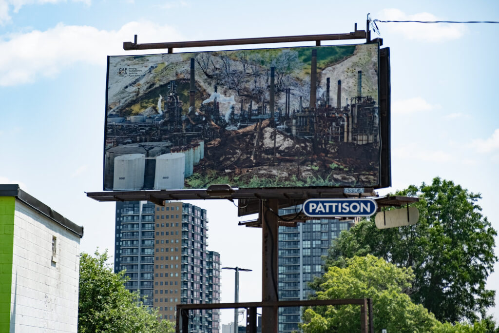 A colour photo of a billboard featuring a photo montage of factory images, smoke stacks, burned trees and a landscape. The billboard is in an urban landscape surrounded by buildings.