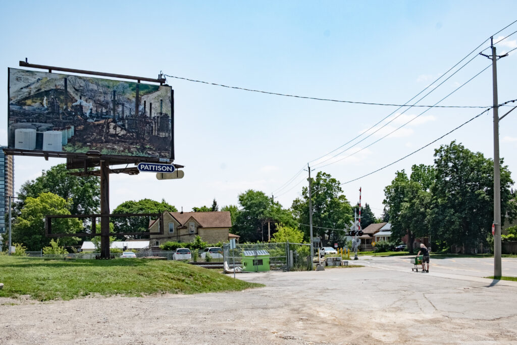 A colour photo of a billboard featuring a photo montage of factory images, smoke stacks, burned trees and a landscape. The billboard is in an urban landscape surrounded by buildings. A man pushing a shopping cart is on the right looking at the billboard from the sidewalk.