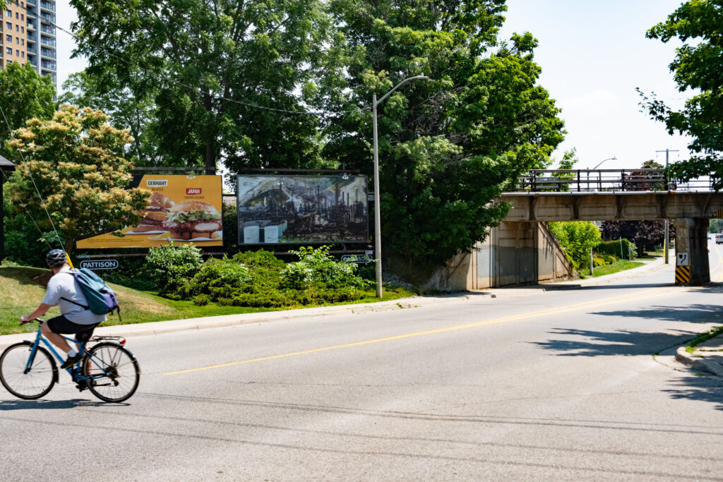 A colour photo of a billboard featuring a photo montage of factory images, smoke stacks, burned trees and a landscape. The billboard is in an urban landscape next to a billboard advertising McDonald's surrounded by shrubbery next to and in front of a railway bridge. A cyclist is riding by in the foreground looking at the billboard.