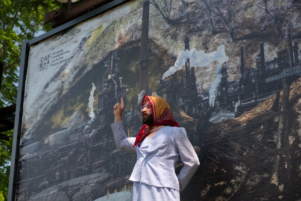 A colour photo of a figure performing in front of a billboard featuring a photo montage of factory images, smoke stacks, burned trees and a landscape.