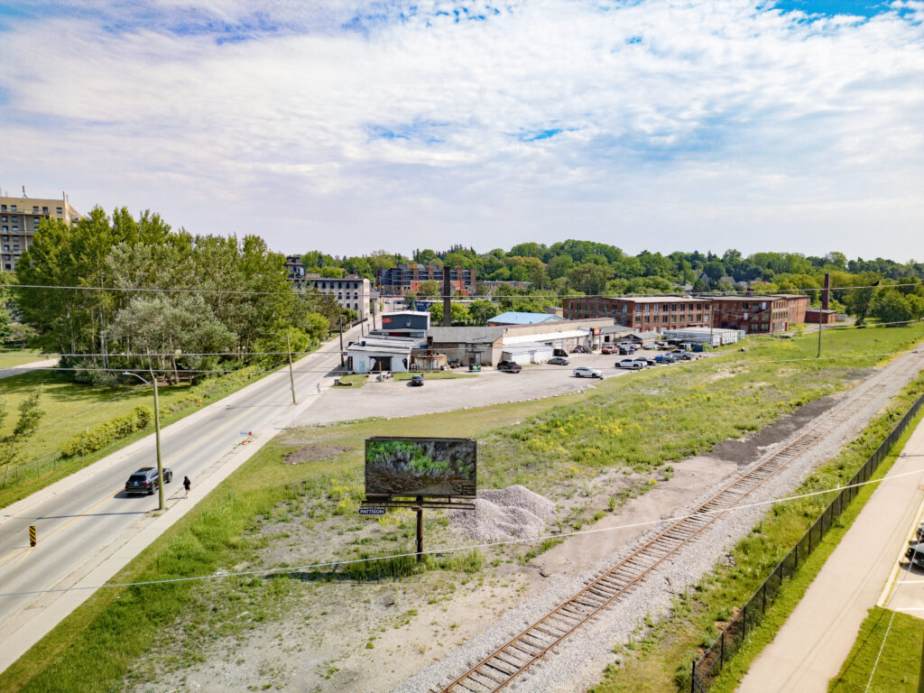 Colour photograph of a Pattison billboard of a photomontage of cut down trees lining a roadway that leads to a forest that has been partially burned. In the foreground under the billboard, is a figure holding a camera in their hands. The billboard is located next to a pile of gravel, in front of old factory buildings and railway tracks run to the right.
