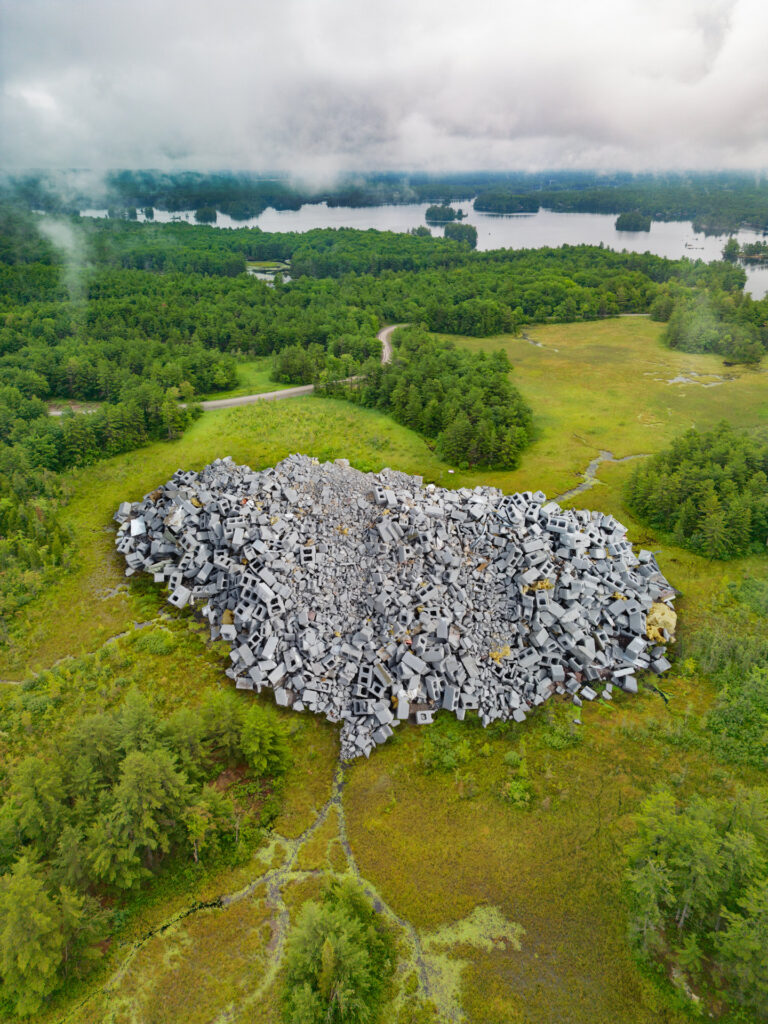 a photomontage of a giant larger than life pile of grey cinderblocks in a misty landscape of connected water ways and forest.