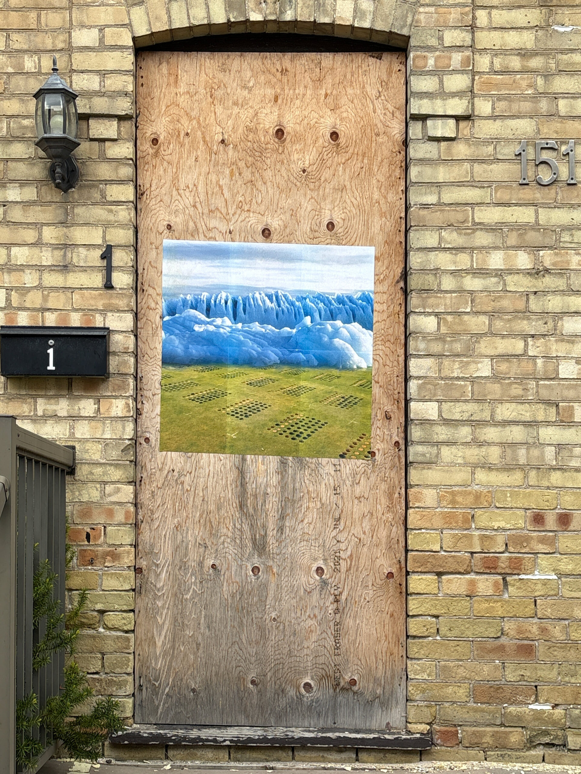 A colour photograph of the front doorway of a yellow brick house, the number 1 is on the left side of the doorway, and the number 151 is on the right. A paste-up of a photomontage of an iceberg and grass is pasted onto the plywood that covers the door.