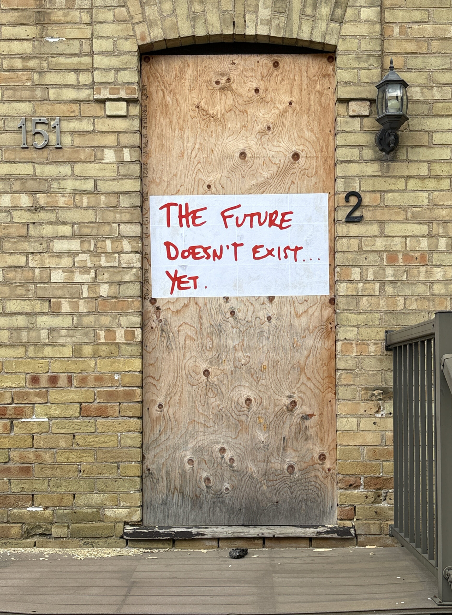 A colour photograph of the front doorway of a yellow brick house, the number 151 is on the left side of the doorway, and the number 2 is on the right. A paste-up of red text that says: The Future Doesn't Exist... on white paper pasted onto the plywood that covers the door.