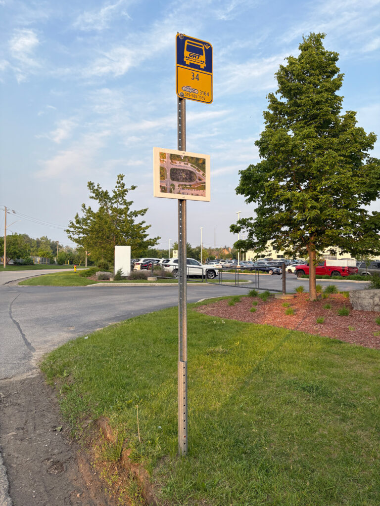 A colour photograph of a yellow and blue coloured bus top sign on a metal post. Below the bus stop is a white plaque and a photomontage of a burned out forest in the center of a photograph of a bus loop aerial image is pasted to it. In the background there is a car dealership parking lot with white and red coloured vehicles and several leafed out trees.
