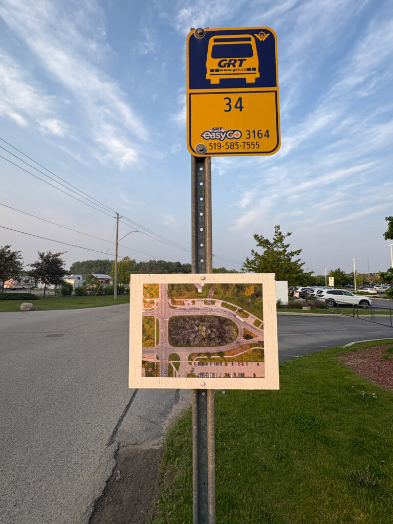 A colour photograph of a yellow and blue coloured bus top sign on a metal post. Below the bus stop is a white plaque and a photomontage of a burned out forest in the center of a photograph of a bus loop aerial image is pasted to it. In the background there is a car dealership parking lot with white and red coloured vehicles and several leafed out trees.