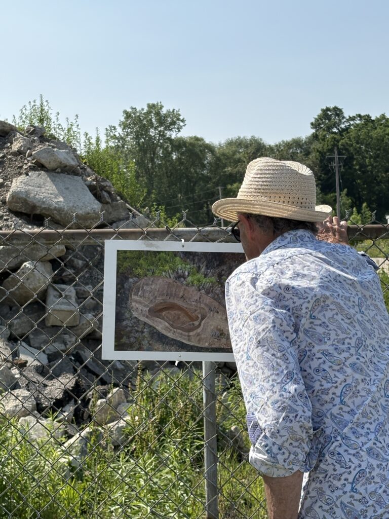 A photograph of a paste up collage that looks like a pit surrounded by burned forest on a white board affixed to a chainlink fence. Behind the fence is a pile of stone rubble. In the foreground a figure in a blue and white long sleeve shirt and straw hat is leaning forward to look at the collage on the board.