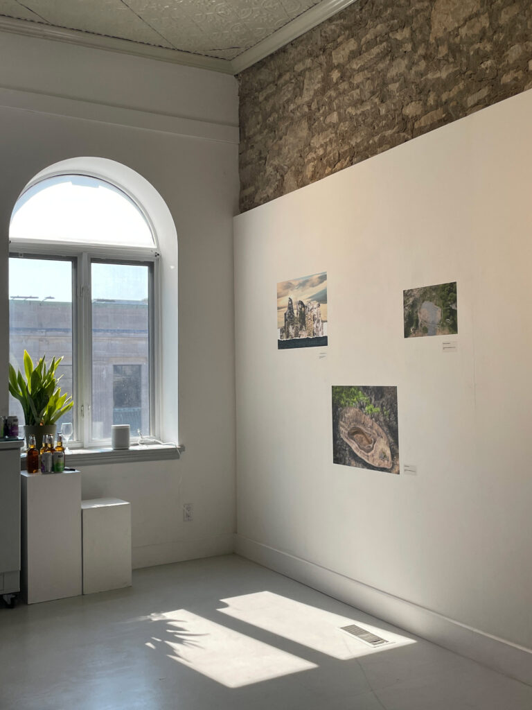A colour photograph of a corner of an art gallery interior, sun is streaming through the window, 3 pieces of art can be seen on a white wall.