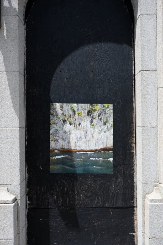A colour photograph of a photomontage of trees and water pasted onto a boarded up doorway painted black.