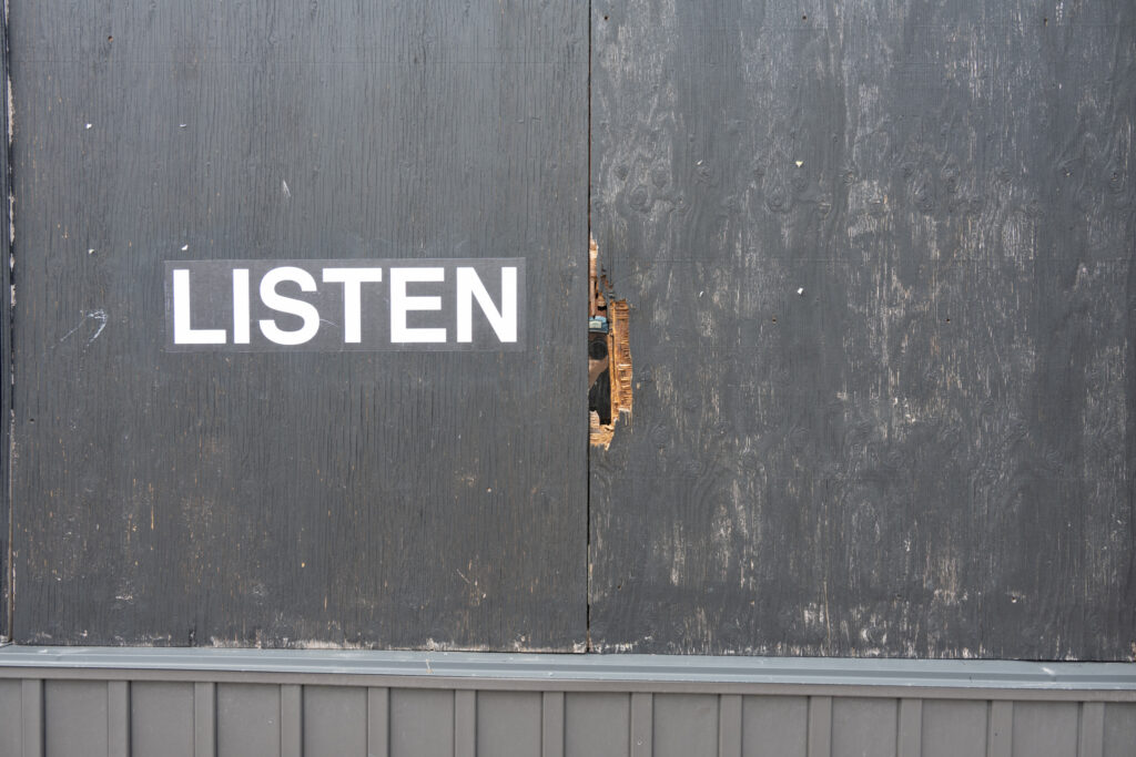 A colour photograph of white text on black paper pasted onto a boarded up surface painted black. In a hole of the black surface you can see a women's face with a camera reflected back.