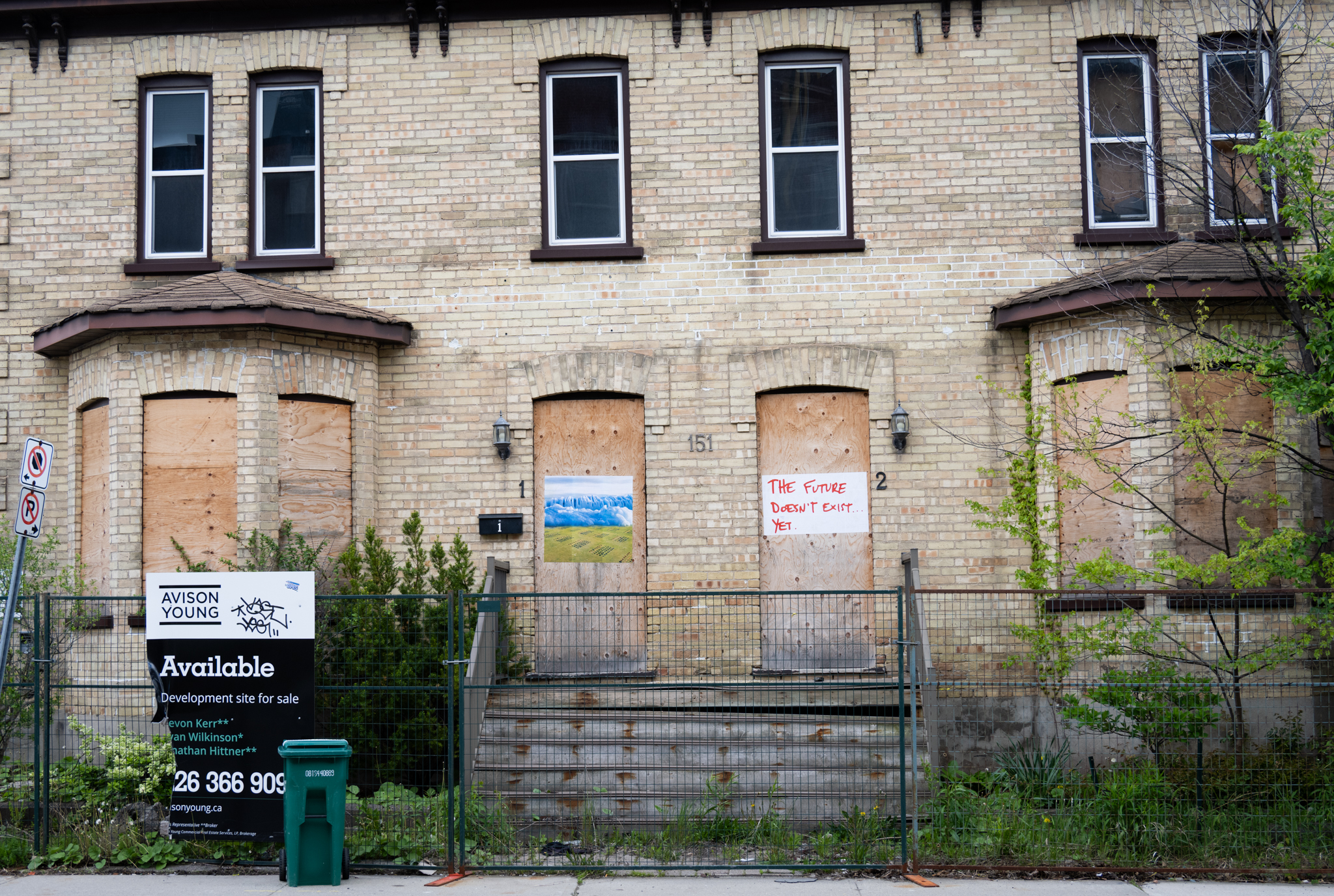 A colour photograph of of a yellow brick duplex house. The ground floor wndows and doors are boarded up with plywood and the house is surrounded by a fence. ON the fence a sign reads "available. A paste-up of a photomontage of an iceberg and grass is pasted onto the plywood that covers the door on the left. A paste-up of red text that says: The Future Doesn't Exist... on white paper is pasted onto the plywood that covers the door on the right.