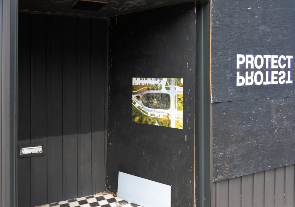 A colour photograph of a photomontage of burned earth and trees pasted onto a boarded up doorway painted black. To the right of the Photomontage are the words PROTECT PROTEST. The word PROTEST upside down and backwards, and acts as a reflection of the word PROTECT.