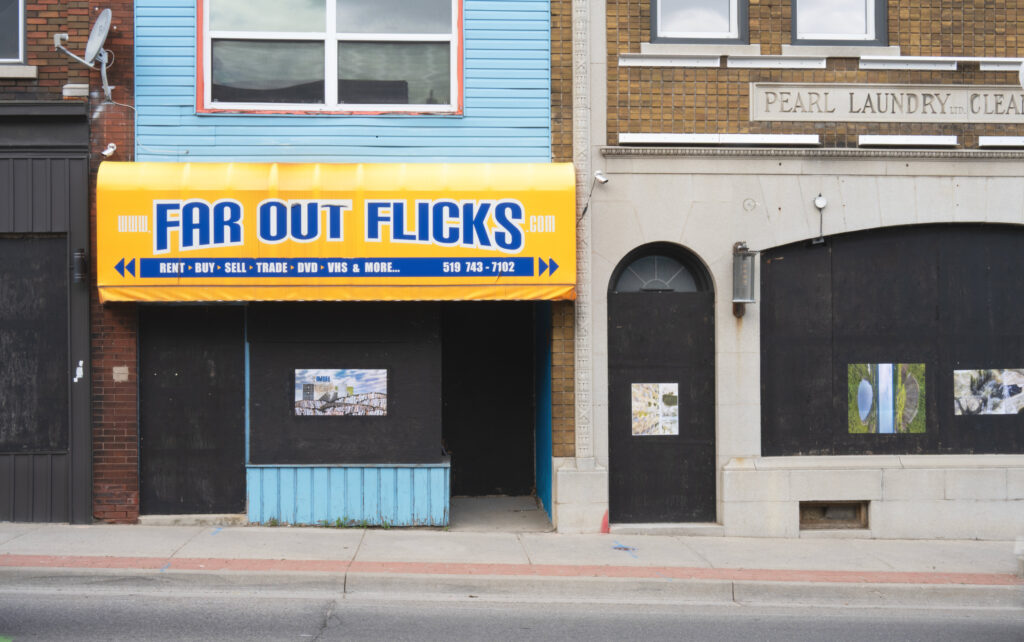 Colour photograph of boarded up storefronts that the coverings of the doors and windows of have been painted black. The building on the left has a yellow awning and blue text that says Far Our Flicks. The building on the right has a brick and cement facade, above the cement archways of the boarde up door and window are the words in relief capital letters: Pearl Laundry Ltd.