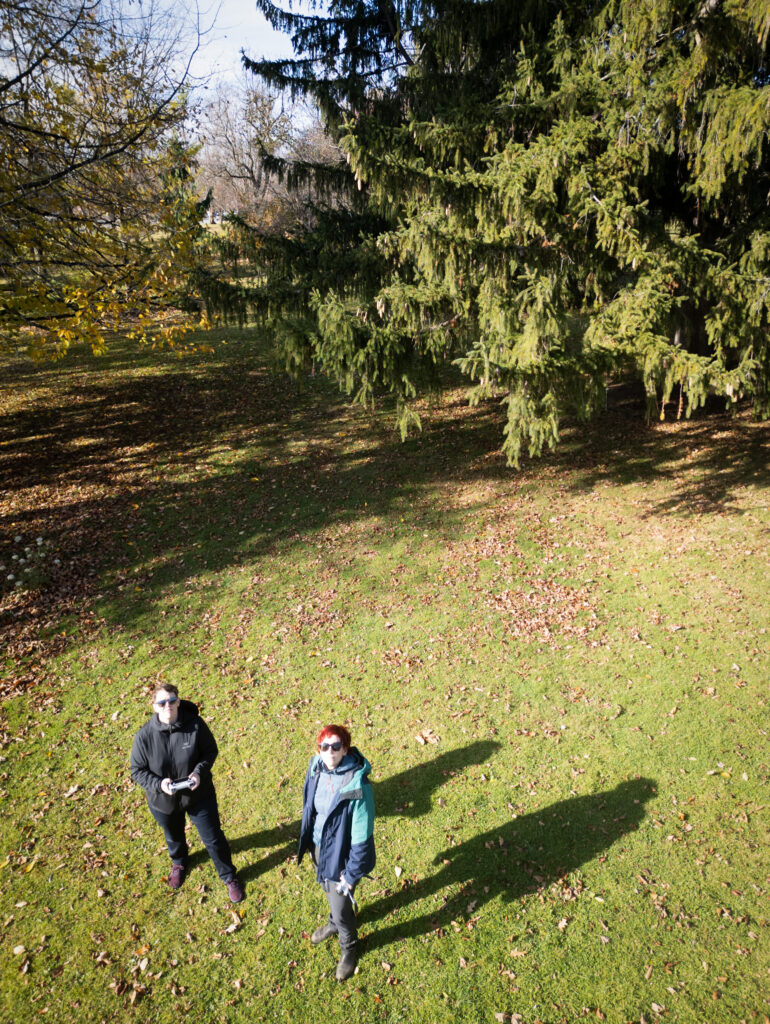 A colour photograph of two women taken from a camera drone. They are looking up at the camera in a park environment. 