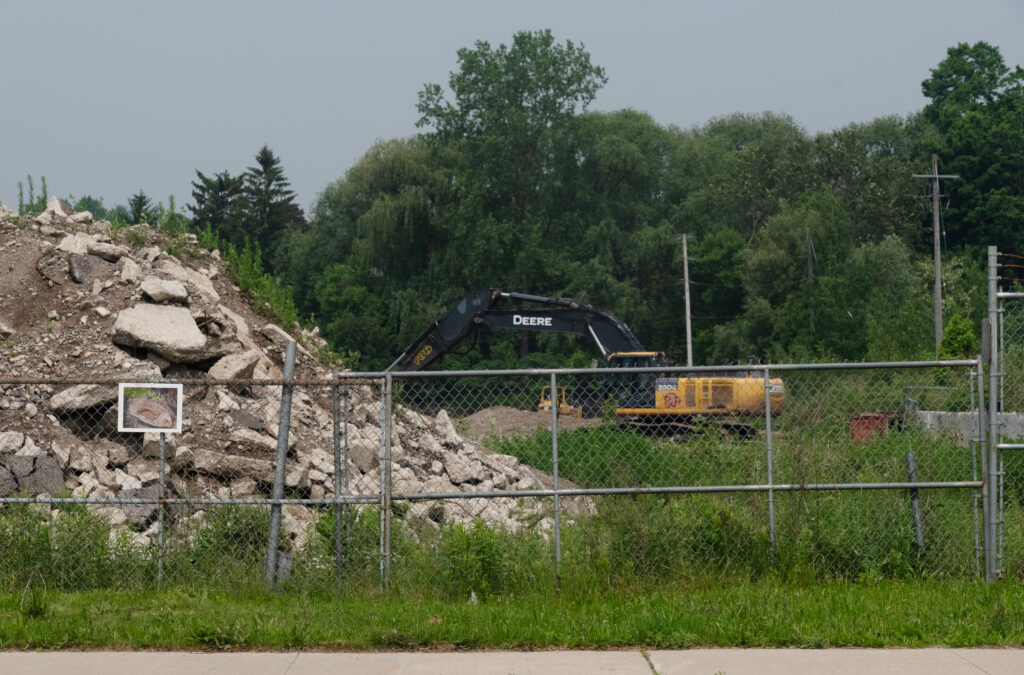 A photograph of a paste up collage that looks like a pit surrounded by burned forest on a white board affixed to a chainlink fence. Behind the fence is a pile of stone rubble and a yellow tractor with a large digging arm.