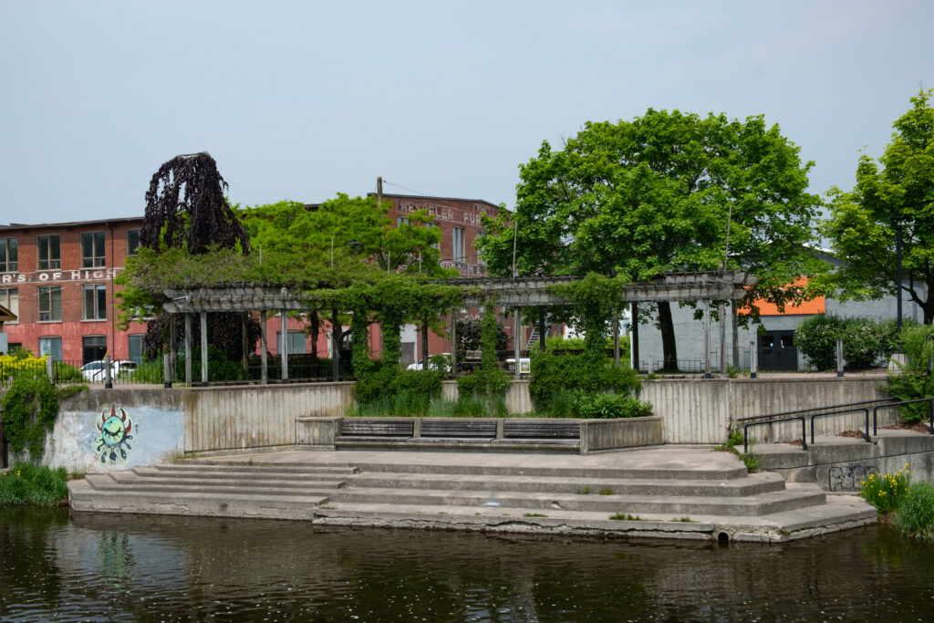 A colour photograph of a concrete sitting area. 3 benches made of wood are on in the center, green plants are growing all around the benches and up a pergola, stairs leading away are on the right, to the left is stone wall with graffit on it. Above the benches is a plaque with a photomontage of a giant larger than life pile of grey cinderblocks in a misty landscape of connected water ways and forest pasted on it. In front of the benches are stairs leading down to a body of water.