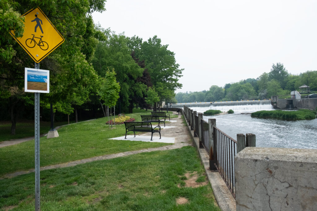 A colour photograph of a line of benches facing a river.
In the foreground on the left is a yellow street sign indicating a walking and cycling route. Below the sign is a white board with a photomontage of photographs of blue ice and a filed of grass growing in pots.
