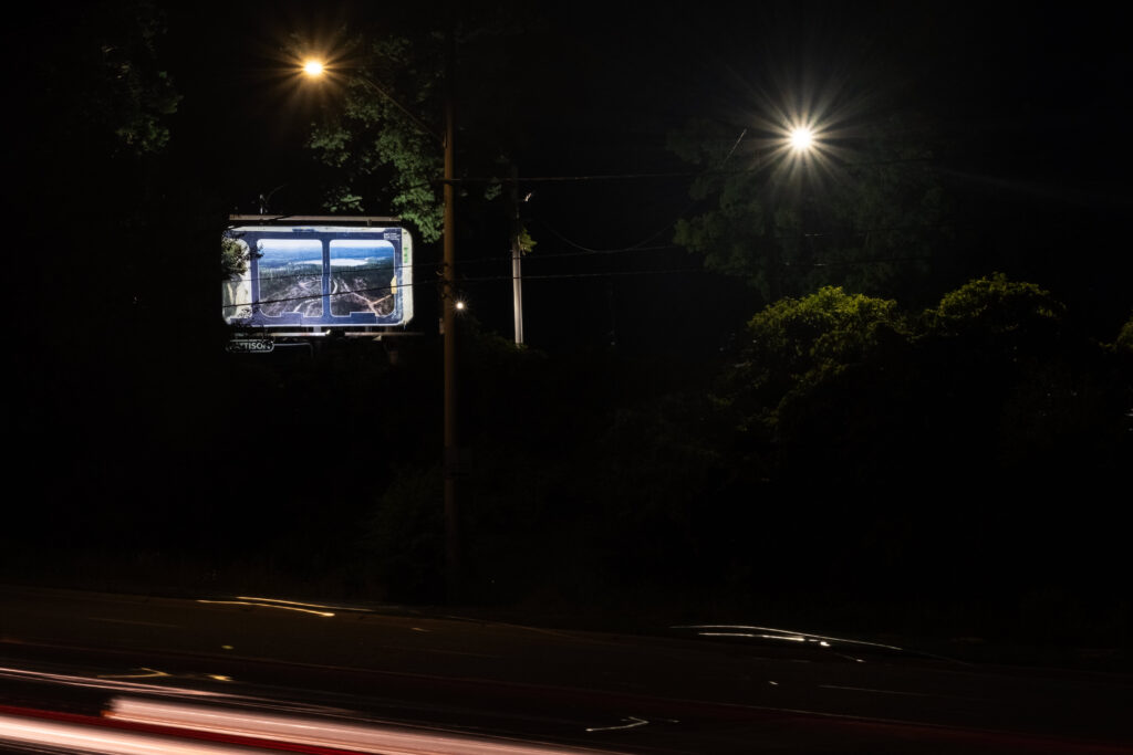 A colour photograph of a Pattison billboard of a photomontage of two photographs. The billboard is surrounded by trees and at the edge of a multi-lane road. It is night, the billboard glows in the darkness.